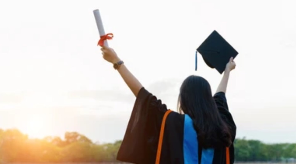 A graduate raising her diploma and cap at sunset.